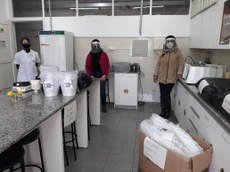 #Pratodosverem: Imagem de uma professora do Campus Barbacena num laboratório de química doando uma caixa de protetores para duas mulheres (uma ao lado da outra). Essas mulheres estão de máscara e protetores faciais.