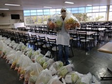 #Pratodosverem: Na imagem, há um nutricionista com jaleco branco, toca e máscara. Ele está segurando duas cestas com frutas e legumes. Há muitas cestas com frutas e alimentos no chão. A imagem é no refeitório.