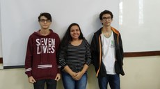 A imagem mostra três alunos lado a lado, sendo dois meninos e uma menina sorrindo, entre eles. Todos estão bem próximos a um quadro branco, no fundo, indicando se encontrarem em sala de aula.