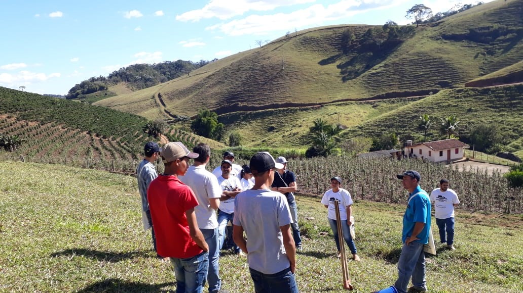 A imagem mostra, em primeiro plano, um grupo de alunos no campo. Ao fundo, uma paisagem composta por plantações, montanhas e uma casa.