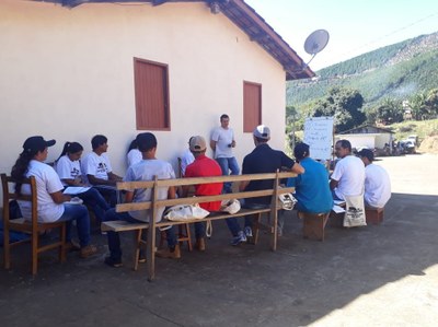 A imagem mostra um grupo de alunos ao ar livre, sentados em bancos de madeira, disposto em formato "U", e voltados a uma espécie de quadro com folhas de papel (flip chart). Próximo ao quadro, há um professor de pé, recostado sobre a parede de uma casa, que proporciona sombra ao grupo.