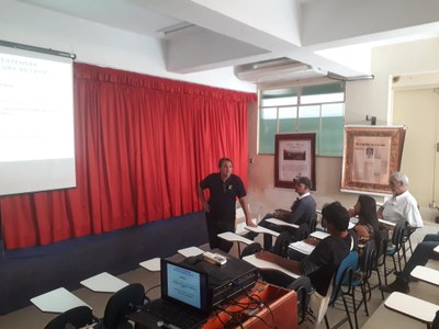 Para todos verem: a imagem mostra uma sala em formato auditório, onde o palco é oculto por cortinas vermelhas. à frente destas, há um painel onde são exibidas, parcialmente, informações sobre o curso de bovinocultura. Quatro pessoas estão sentadas de frente para o palco/ painel, enquanto um professor do IF Sudeste MG está posicionado de pé, voltado tanto para os presentes, quanto para o painel em exibição.