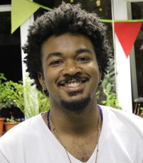 #pratodosverem: a imagem é uma fotografia de rosto do professor convidado, Diego Dhermani. Ele usa uma camiseta branca e aparece sorrindo. Ao fundo, há plantas e bandeirolas coloridas.