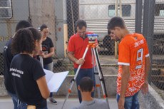 #pratodosverem: a imagem é uma fotografia de uma aula prática de Engenharia Ferroviária e Metroviária no Laboratório de Manutenção Ferroviária, no Campus Santos Dumont. Aparecem alguns alunos de camiseta preta do lado esquerdo e um aluno de camiseta laranja, do lado direito. Ao centro, o professor Fernando Caneschi manuseia um equipamento apoiado em um tripé.