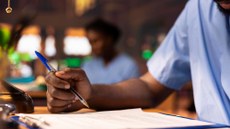 Imagem de uma pessoa preta de camisa azul, desfocada, ao fundo. Em primeiro plano, o braço de uma pessoa preta, com uma caneta de tampa azul escrevendo em um papel.