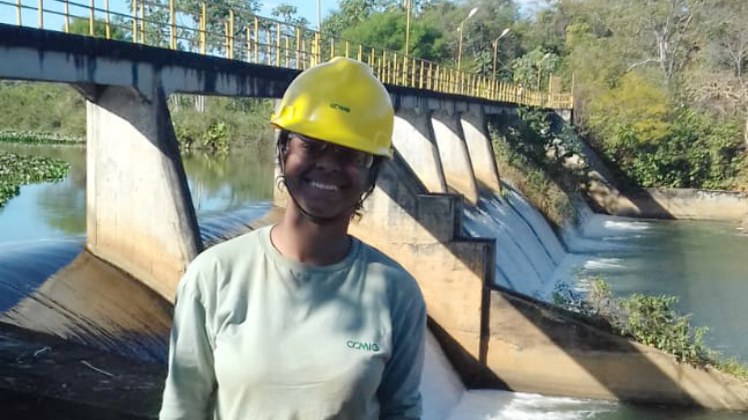 A aluna do Campus está na Usina de Pandeiros. Ela aparece em primeiro plano, usando um uniforme cinza e um capacete amarelo. Ao fundo, uma passarela construída em cima das águas, que caem em forma de cachoeira.