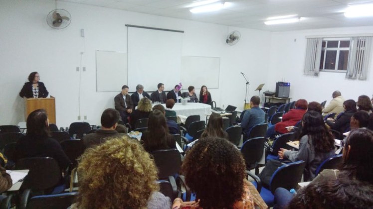 Aula inaugural de 2018 Foto sobre a aula inaugural do curso ProfEPT em 2018. Ao fundo da imagem, uma mesa formada pelos membros da coordenação do curso. Ao lado esquerdo, a apresentadora do evento em pé em frente a um púlpito. No primeiro plano da foto, os alunos sentados em cadeiras azuis de frente para a mesa.