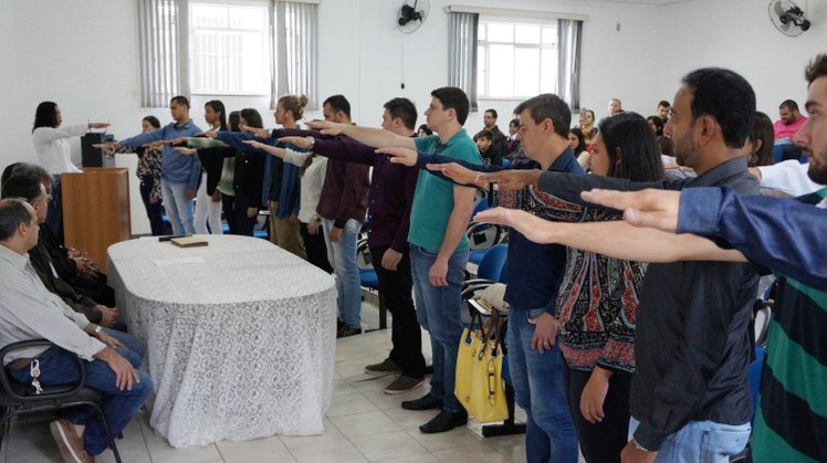 Foto / Arquivo. Imagem retrata colação de grau em gabinete realizada em 2018. Ao fundo da imagem, uma estudante de blusa branca está no púlpito, proferindo o juramento. À sua frente, os demais formandos de pé e com a mão direita à frente. Ao lado dela, equipe de direção sentada à mesa.