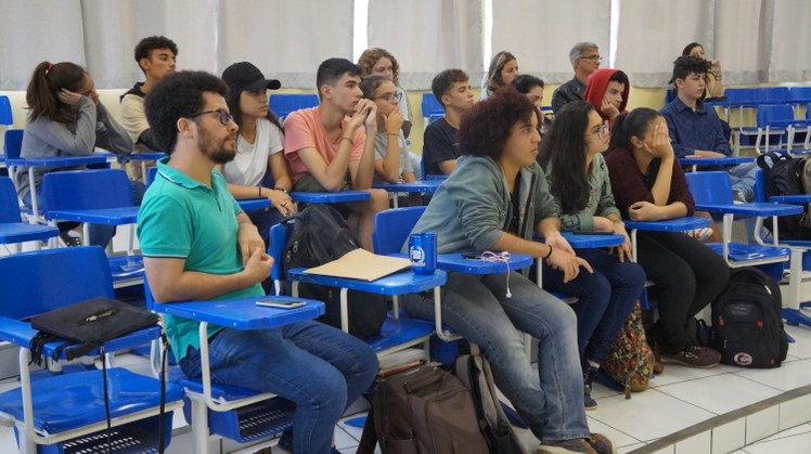 Vários alunos sentados em carteiras azuis, olhando fixamente para a frente.