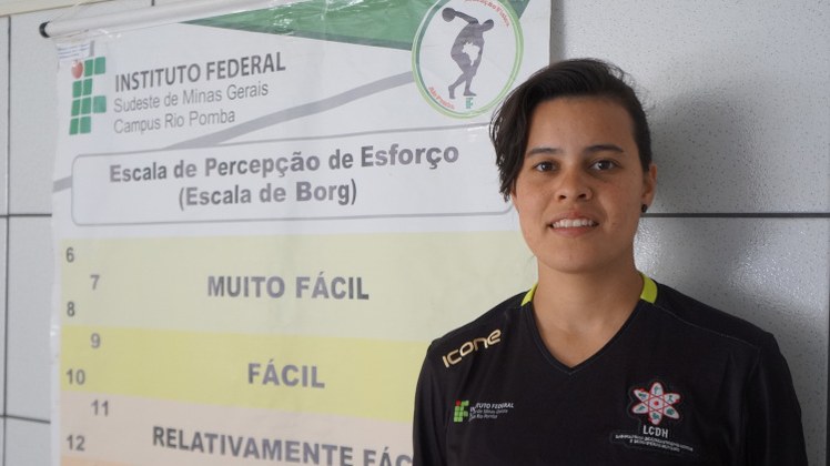 Suene Chaves A aluna Suene Chaves está de camisa preta com o símbolo do IF. Ao lado, um pôster com o título "Escala de percepção de esforço (escala de Borg).
