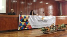 Ao centro da imagem, aparece uma mesa com uma faixa alusiva ao evento. À frente, há um arranjo de flores em tons de alaranjado. Atrás, dois palestrantes sentados. Um deles o estudante do Campus Rio Pomba.