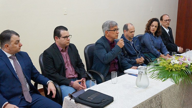 Aula Magna Direito Cinco pessoas formando a Mesa de Abertura. O diretor-geral João Batista Lúcio Corrêa, ao centro da imagem, segura um microfone. A mesa tem toalha branca e arranjo de flores.