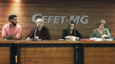 Foto: Congresso Teias
Em primeiro plano, uma mesa de madeira. Quatro pessoas sentadas atrás, incluindo as duas professoras do Campus. Ao fundo, uma parede em madeira com o letreiro Cefet - MG