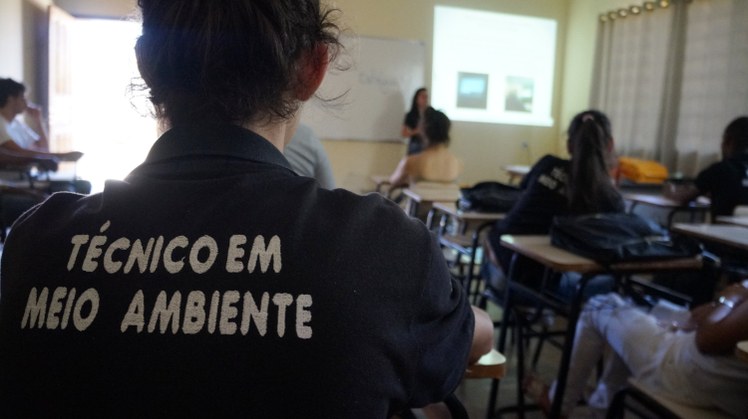 #Pratodosverem: Um estudante sentado de costas. Em sua camisa, está escrito "Técnico em Meio Ambiente". Ao lado dele, pessoas sentadas acompanhando apresentação. Ao fundo, uma pessoa em pé falando ao lado de uma projeção de fotos.
