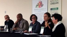 #Pratodosverem: Elisete Reis está sentada em mesa ao lado de demais participantes do evento. Em cima da mesa, que está com toalha preta, há copos e jarra de água. Ao fundo, dois pôsteres estão na parede.