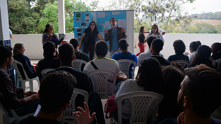 #Pratodosverem: os estudantes surdos e a professora Bárbara ensinam sinais à plateia. Ao fundo, há um quadro com as palavras a serem aprendidas. Em primeiro plano, estão os estudantes sentados em cadeiras e fazendo os sinais. Ao fundo, há árvores.