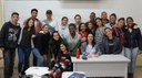 #Pratodosverem: estudantes de Meio Ambiente participantes da Olimpíada. O grupo está em pé em frente a um quadro branco. A professora Flávia está sentada atrás de uma mesa que tem uma garrafa alaranjada em cima. Ao lado dela, duas alunas agachadas.