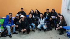 #Pratodosverem: estudantes de Agropecuária participantes da Olimpíada. Seis alunos estão sentados em carteiras e os demais agachados, assim como a professora. Ao fundo, uma parede alaranjada.