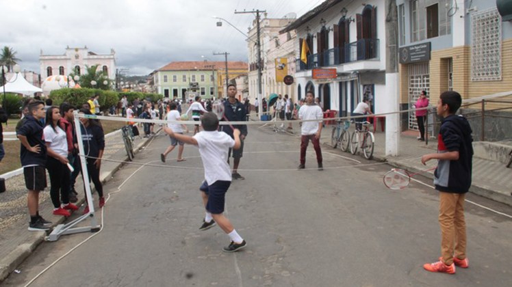 Educação Física na Fliminas #Pratodosverem: Crianças jogam badminton em quadra improvisada no meio da rua. Há pessoas de pé assistindo a partida. A área da praça tem vários casarões antigos.