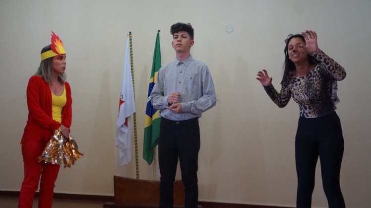 Apresentação teatral #Pratodosverem: À esquerda, está a personagem Fogo. Ela veste roupas vermelhas e amarelas. Usa o desenho de uma chama na cabeça. Ao centro, está o juiz. Ele usa camisa e calça social. À direita, está a Onça. A atriz está com uma camisa de estampa de onça pintada e uma calça preta. No rosto, desenhou os bigodes do animal.