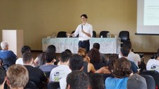 #Pratodosverem: O palestrante está de pé, olhando e gesticulando em direção ao público. Ele segura um microfone. À sua frente, várias pessoas sentadas. Ao fundo, uma mesa com toalha de renda branca e cadeiras. Em cima da mesa, jarras com água e copos. À esquerda, há um púlpito. À direita, uma tela de projeção.