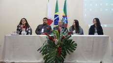 #Pratodosverem: Mesa de abertura do evento. Cinco pessoas compõem a mesa. A da esquerda está com microfone na mão. Ao fundo, há bandeiras em mastros. À frente, um arranjo de flores.