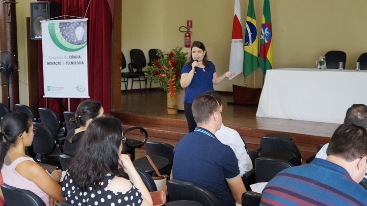 Simpósio de Ciência, Inovação e Tecnologia #Pratodosverem: Aluna faz apresentação oral de seu trabalho no Salão Nobre. Ela está à frente da plateia. Ao lado dela, há banner do evento.