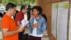 #Pratodosverem: Aluna apresenta pôster para o professor. Á direita, está o poster. À esquerda, o avaliador com papel e caneta na mão. Ao fundo, há outras pessoas.