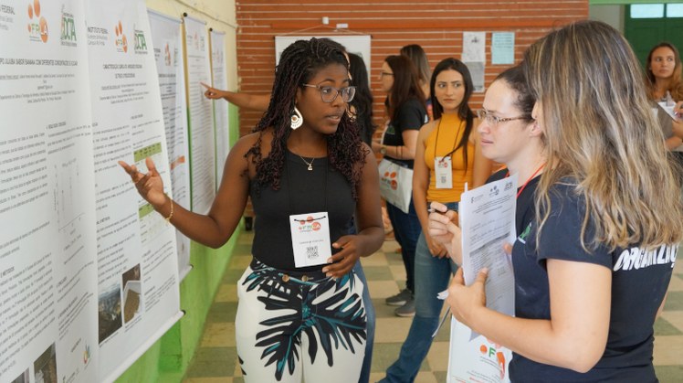#Pratodosverem: Participantes do evento apresentam trabalhos em forma de pôster. Em destaque, estudante explica seu trabalho para outras duas pessoas. Ela aponta para o pôster.