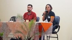 #Pratodosverem: Palestrante e membro da organização estão sentados à mesa. A mulher, que está à direita, fala ao microfone. A mesa tem toalha de alaranjada no fundo em, que é a cor do evento, e uma de renda branca por cima. Há folhagens à frente da mesa.