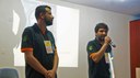 #Pratodosverem: Organizadores do evento fazem abertura oficial. Os dois estão com a camisa do evento em preto e alaranjado. O da direita está com microfone na mão. Ao fundo, há projeção.