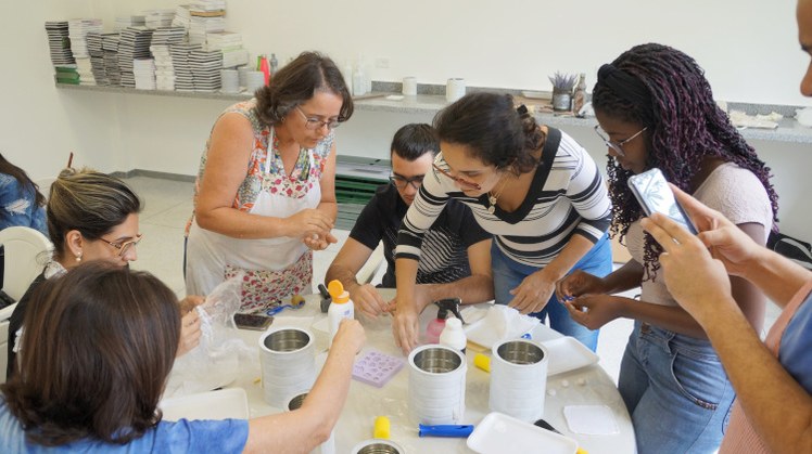 #Pratodosverem: Estudantes e servidores participam de oficina de artesanato. Eles estão em volta de uma mesa, alguns em pé e outros sentados, pintando latas.