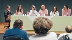 #Pratodosverem: Seis pessoas compõem a mesa de produtos educacionais. Camila é a segunda da esquerda para a direita. Ela segura um microfone. Outros três participantes aparecem em primeiro plano de costas. Eles acompanham a apresentação.