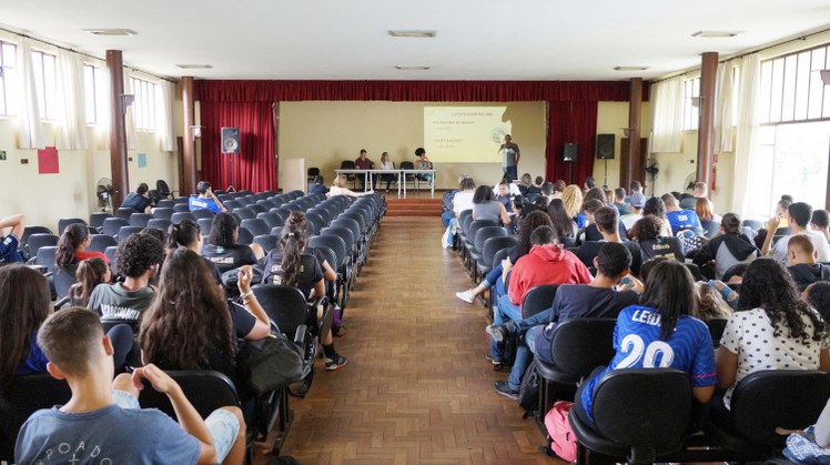 #Pratodosverem: Imagem do Salão Nobre durante o evento. Ao centro, há um corredor. Ao lado, cadeiras ocupadas por estudantes. Ao fundo, aparece o palco e os palestrantes.