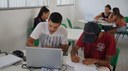 #Pratodosverem: dois estudantes aparecem em primeiro plano, sentados. Há uma mesa na frente deles com computador, caderno, calculadora, estojo e papéis avulsos. Atrás de um dos meninos, há uma aluna mexendo em um computador. No fundo, outras duas alunas conversam e uma delas mexe no notebook.                               