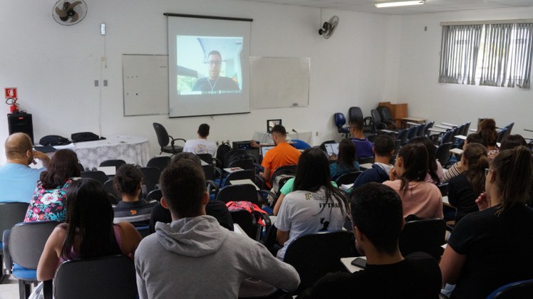 #Pratodosverem: Estudantes acompanham estão sentados no anfiteatro, acompanhando a transmissão. Na parte da frente da sala, há a projeção do palestrante.