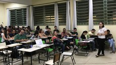 #Pratodosverem: Sala de aula lotada. Alunos e professores estão sentados nas carteiras, olhando em direção à parte da frente da sala, onde uma pessoa faz apresentação.