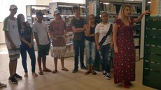 #Pratodosverem: A gerente de Acervos Bibliográficos e Multimeios, Tatiana dos Reis, está de pé ao lado das estantes com livros. Ela explica aos alunos que estão ao lado como localizar um livro no acervo.