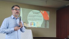 #Pratodosverem: O pró-reitor de Pesquisa, André Narvaes, aparece em primeiro plano com microfone na mão. Ao fundo, há a projeção da apresentação de sua palestra