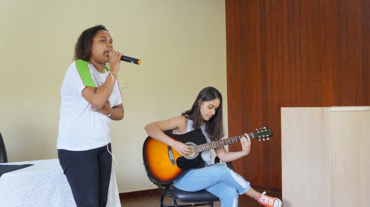 Dia da Mulher #Pratodosverem: Estudantes fazem apresentação musical. Uma está de pé com microfone na mão. A outra sentada, tocando violão.