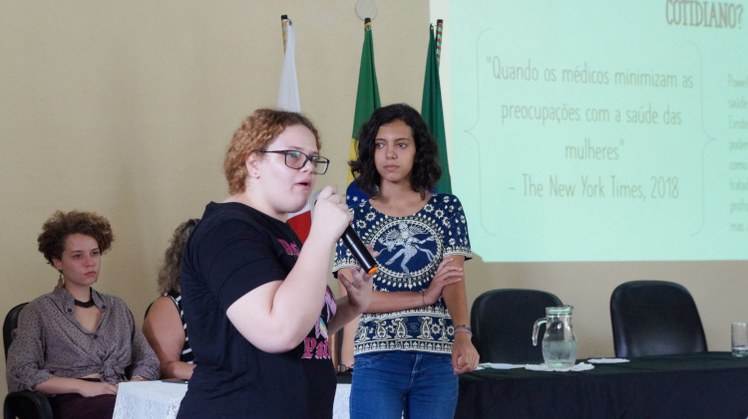 Dia da Mulher #Pratodosverem: Alunas fazem apresentação. Uma delas está com microfone na mão.