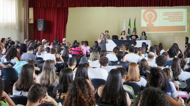 Dia da Mulher #Pratodosverem: Professora faz palestra no Salão Nobre para plateia de alunos. No palco, há mesa composta pelas palestrantes e, ao fundo, projeção da apresentação.
