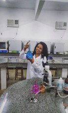 #pratodosverem: em um ambiente laboratorial fechado, composto por balcões, tubos de ensaio, microscópio e outros, Heloísa Mendes sorri, vestida de branco, enquanto segura um tubo de ensaio azulado.