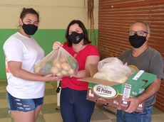 #pratodosverem: a imagem é uma fotografia onde aparecem três pessoas. Uma jovem de camisa branca, à esquerda, aparece recebendo uma sacola e uma caixa entregues por servidores localizados à direita da imagem. Todos usam máscaras.