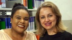 #pratodosverem: Imagem mostra bibliotecárias Paula Souza e Érica Veloso. Elas estão sorridentes. Ao fundo, aparece uma estante com livros na Biblioteca Acyr Loureiro Lima.                               