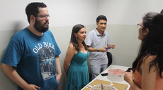 #pratodosverem: Imagem mostra evento da Licenciatura em Matemática realizado em 2019 no Campus Santos Dumont. Três estudantes do curso apresentam um jogo didático à professora Francilene Barbosa.