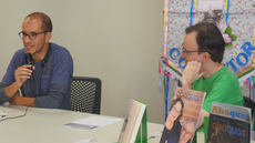 #pratodosverem: Imagem mostra músico Tiago Guimarães e diretor-geral Benedito Carvalho conversando. Tiago fala ao microfone. Eles estão sentados a uma mesa com exemplares da revista Abaquar e do livro "Contos da Cidade de Santos Dumont II".