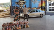 #pratodosverem: Imagem mostra, à esquerda, representante do Coletivo Manas pelas Minas e, à direita, um dos integrantes da Subcomissão de Enfrentamento á Covid-19 do Campus Santos Dumont, professor Carlos Artur Leal. À frente, estão dez cestas básicas com alimentos e kits de higiene, destinadas a mulheres chefes de famílias em vulnerabilidade social durante a pandemia. Ao fundo, aparece o painel artístico em homenagem ao Pai da Aviação, localizado no Bloco 1 do Campus Santos Dumont.