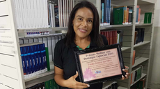 #pratodosverem: Imagem mostra bibliotecária Paula Souza com a placa do "Prêmio Mulher em Destaque". Atrás dela está uma estante da Biblioteca Acyr Loureiro Lima.