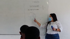 #Pratodosverem: Imagem mostra residente Juliana Vitorelli, da Licenciatura em Matemática, explicando frações durante uma aula na Escola Estadual Engenheiro Henrique Dumont. Aparecem na imagem dois alunos, de frente para a residente, observando o quadro atentamente.